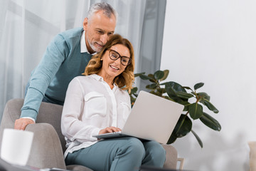 attractive woman in eyeglasses using laptop while her mature husband standing behind at home