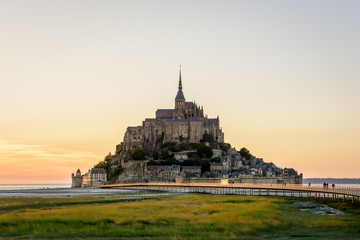 View of the Mont Saint-Michel tidal island at sunset and low tide with a shuttle bus driving on the jetty on stilts, people strolling on the walkway and salt meadows in the foreground.