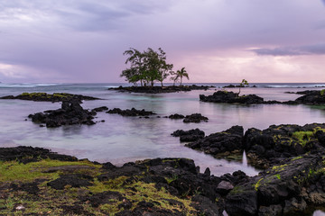 Rocky shore at sunrise in Hilo, Hawaii. Volcanic rock and grass on shore; crystal clear water with rocks at low tide; trees on offshore outcrop. Pacific ocean in distance, storm clouds overhead.
