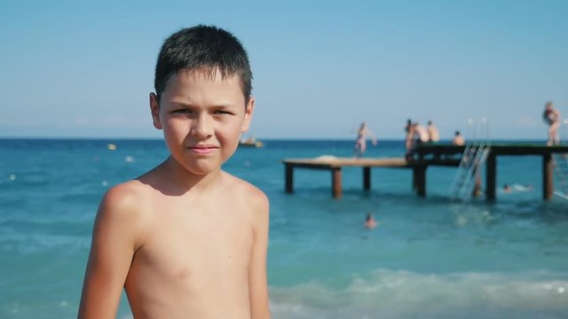 Midshot of a funny small boy with a big smile and screwed up eyes standing on sea beach with a wooden platform behind him in summer in slow motion