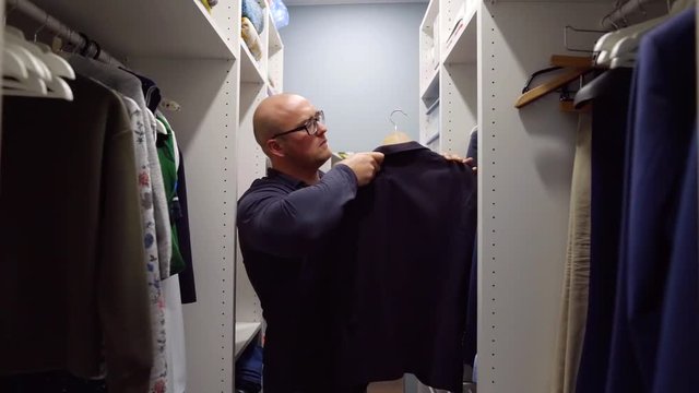 bald man is taking hangers with jackets and shirts in wardrobe room, deciding what to wear