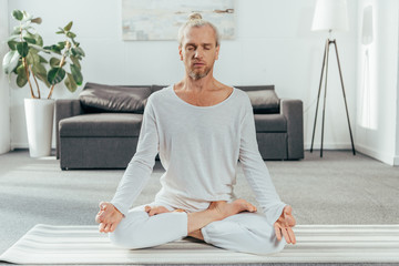 sporty adult man meditating in lotus position on yoga mat at home - obrazy, fototapety, plakaty
