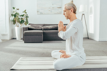 side view of man meditating in lotus position with namaste gesture at home