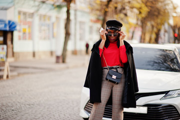 African american fashion girl in coat, newsboy cap and sunglasses posed at street against white business car.