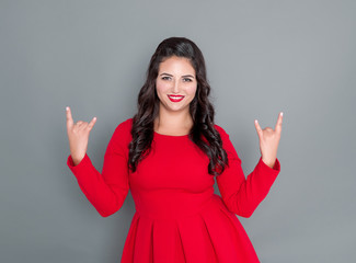 Happy plus size woman in red dress showing horns gesture on gray