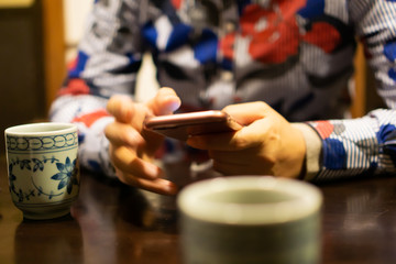 Fototapeta na wymiar Hands of young woman using smartphone in traditional Japanese restaurant, with ceramic cup of Japanese roasted green tea in the foreground (This tea looks like Chinese oolong tea)
