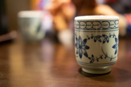 White ceramic cup of Japanese roasted green tea on a table in traditional Japanese restaurant, with young asian woman using smartphone and waiting for her food in the background (The tea is called Hoj