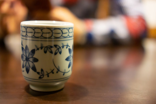 White ceramic cup of Japanese roasted green tea on a table in traditional Japanese restaurant, with young asian woman using smartphone and waiting for her food in the background (The tea is called Hoj