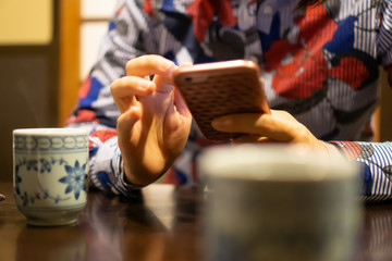 Hands of young woman using smartphone in traditional Japanese restaurant, with ceramic cup of Japanese roasted green tea in the foreground (This tea looks like Chinese oolong tea)