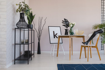 Black mannequin's leg on wooden chair in elegant dining room interior with copy space on the empty wall, flowers and leaf in vases on the floor and poster on the chair