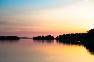 The beautifully reflected pastel hues of sunset a few days after midsummer, viewed through a backdrop of trees and clouds in a cove on the Island of Nicklösa in the Åland Islands, Finland.