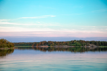A beautiful pastel blue sunset a few days after midsummer, with a tree-lined horizon and clouds reflected on the water of a cove on the Island of Nicklösa in the Åland Islands, Finland.