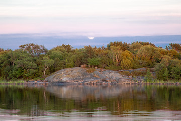 The moon gently rises in the distance behind trees and a rocky shoreline in a cove on the Island of Nicklösa in the Åland Islands, Finland, at sunset a few days after midsummer.