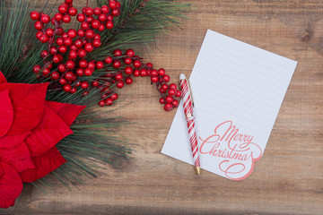 Winter Holidays decoration red poinsettia, pine and berry bush with white Merry Christmas note and red and white striped pen on wooden background.