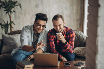 Romantic partners in love and business. Caucasian amused gay couple looking at laptop screen while resting in living room with loft interior. Dominant man wearing spectacles and and using laptop - Powered by Adobe