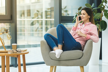 Enjoying time at home. Beautiful young smiling woman using phone and drinking coffee while sitting in a big comfortable chair at home. Large copyspace.