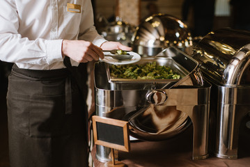 A waiter with tongs take a vegetables fron the bowl on the buffet table