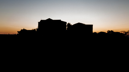 Silhouette of two houses isolated on top of a mountain, while behind them the sun is shining, coloring the sky with red and orange.