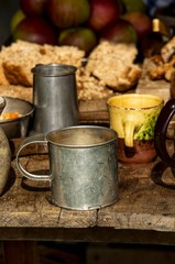 Pewter cups and bowls on a table, hunks of bread and apples in the background.