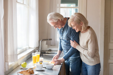 Smiling aged wife hug man from behind watching him preparing healthy food, loving senior woman embrace husband cooking breakfast slicing fruit, romantic couple spend morning at home together - obrazy, fototapety, plakaty