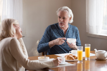Happy senior couple enjoy morning at home eating healthy breakfast, having coffee, smiling aged husband and wife talk at kitchen table spending weekend together, elderly man and woman chat laughing