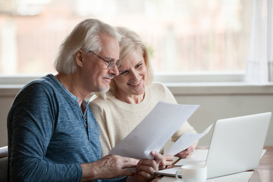 Happy aged husband and wife hold papers using laptop for online banking, satisfied senior couple smiling checking utility bills or insurance at computer with easy access, elderly users of technology