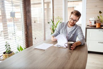 Cheerful male landlord sitting by desk in the kitchen and looking at lease contract at well lit and...