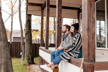 Happy young couple sitting outdoor, in front of their new home in autumn warm clothes and dreaming...