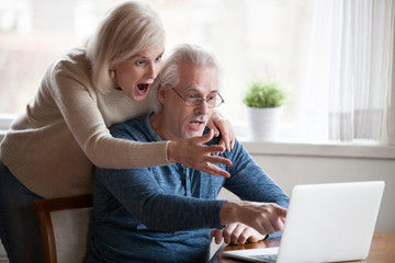 Shocked senior husband and wife pointing at laptop screen seeing unbelievable information,...