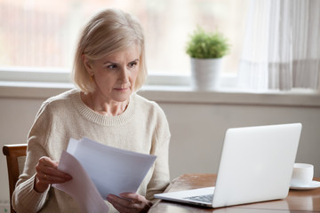 Serious aged woman holding documents, checking information at laptop online, concerned senior...