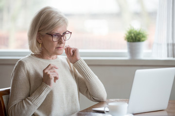 Surprised aged woman using laptop worried seeing error message on screen, concerned senior female...