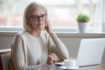 Upset aged woman using laptop at home suffering from headache, tired senior female massaging...
