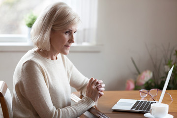 Thoughtful aged woman working at laptop thinking or considering something, doubtful senior female...