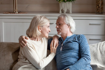 Happy senior husband and wife look at each other talking relaxing on couch, smiling aged couple cuddle and embrace on sofa at home, romantic elderly man and woman enjoy rest having conversation