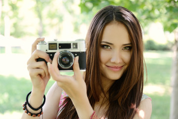 Cheerful beautiful girl talking pictures with vintage camera in summer park. Young woman photographer posing with her camera outdoor.