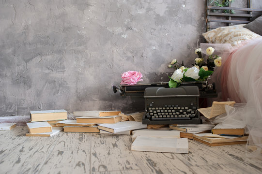 Closeup photo of a vintage typewriter covered with white and pink flowers on a wooden floor with white books on it close to curtains near a window