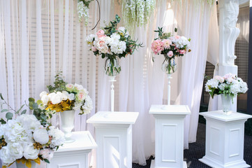 Closeup photo of white pillars with vases with flowers on them and a part of an wedding arch with white curtains on background