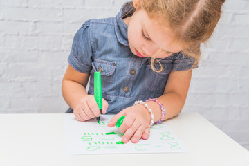 a child, a girl at the table writes, draws on a piece of paper, against a white brick wall