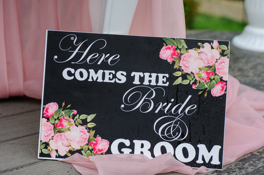 A here comes the bride and groom inscription with white letters on a black background with flowers, on the floor near a table with a pink curtain