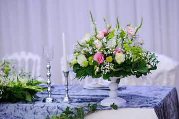 Wedding presidium table with white and grey tablecloth, floral arrangements: a bouquet o flower in a white vase, candles in silver support, white chair on background