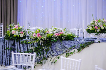 Wedding presidium table with white and grey tablecloth, floral arrangements: a bouquet o flower in a white vase, candles in silver support, white chairs and gray curtains on background