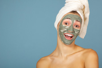 Beautiful cheerful teen girl applying facial clay mask. Beauty treatments, isolated on blue background.