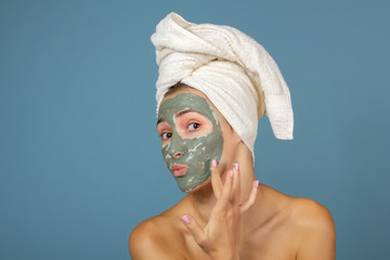 Beautiful cheerful teen girl applying facial clay mask. Beauty treatments, isolated on blue...