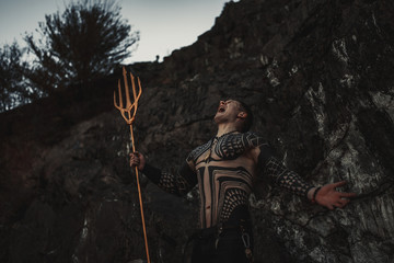 A fury man with a trident in his hands against the background of rocks.