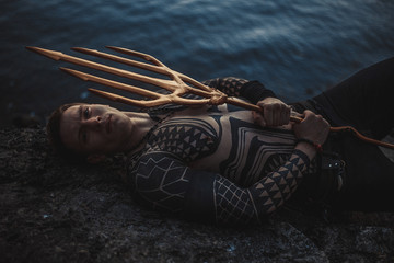 A young man with a trident in his hands lies on the rock near water. - obrazy, fototapety, plakaty