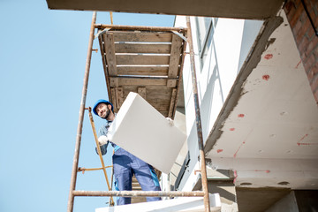 Builder warming a building facade with foam panels standing on the scaffoldings on the construction site