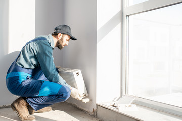 Workman mounting water heating radiator near the window in the white renovated living room, Image...