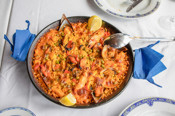 above view of typical Spanish paella with seafood (mussel, prawn, lobster, squid, clam), rice and lemon ready to serve on white tablecloth of restaurant