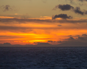 Naklejka na ściany i meble Brilliant orange and yellow sunset at sea with mountains and clouds in silhouette