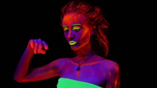 A beautiful young sexy half-naked girl dancing with glowing paint on her body in black light. Pretty woman with glowing bodyart in black lamp light. Waving hands, smiling and beckoning.